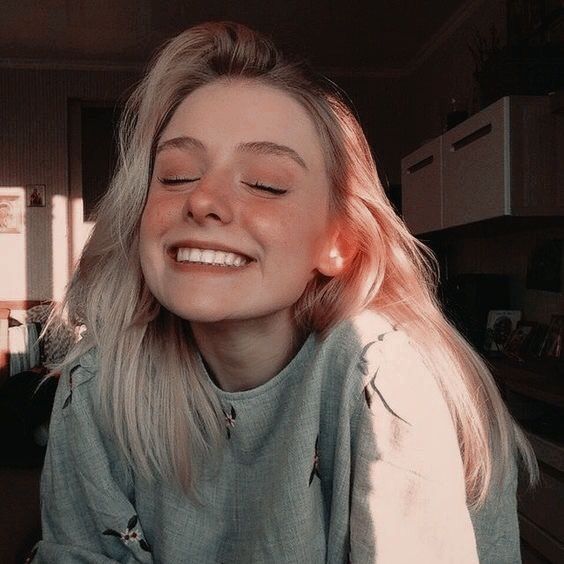 a young blonde woman with rosy cheeks is smiling broadly, her eyes closed in enjoyment. she is wearing a light grey sweater. the image is taken from a closeup angle, and the woman's face is the main subject. the background is blurred, and the light is soft and warm, creating a warm and inviting feel.