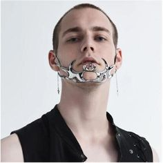 a closeup shot of a man with a shaved head and a silver metallic beard jewelry. the jewelry features a flamelike design with intricate details and a small, round pendant in the center. the man is wearing a black sleeveless shirt. the background is a neutral gray, allowing the jewelry to stand out prominently. the image is welllit, highlighting the shine and texture of the metal.