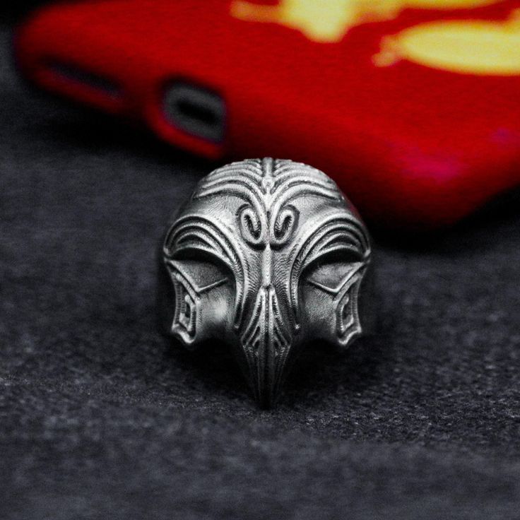 a closeup shot focuses on a silver ring, intricately carved with a detailed face design, placed on a textured dark surface. in the blurred background, a red phone case is visible, adding a splash of color to the composition. the ring appears antique or vintage, its metal displaying complex patterns and shapes that form the face.