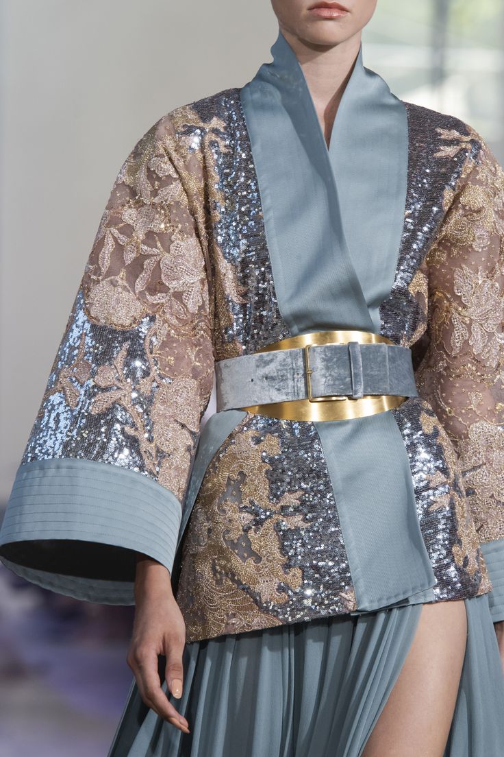 closeup view of a model showcasing a haute couture ensemble. the outfit consists of a light blue kimonostyle jacket adorned with silver sequins and gold embroidery.  a wide, dark blue velvet belt with a gold buckle cinches the waist. the jacket is worn over a pleated skirt in a similar shade of blue. the overall style is elegant and sophisticated. the image focuses on the intricate details of the fabric and embellishments.