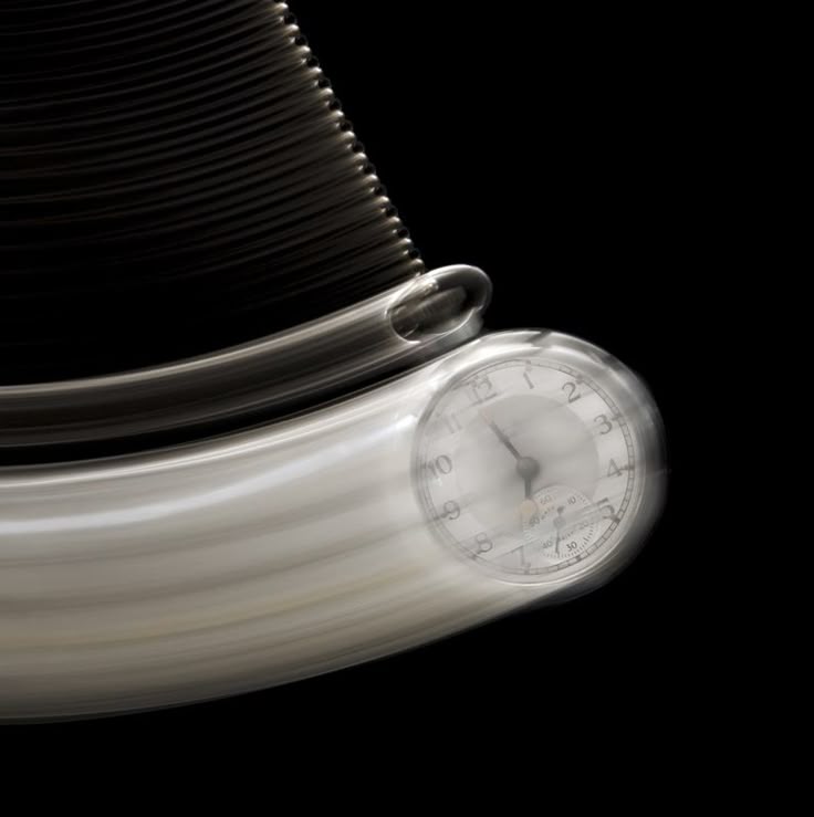 the image shows a silver pocket watch attached to a pendulum, swinging against a black background. the movement is captured with motion blur, creating streaks that emphasize the speed and arc of the pendulum. the face of the watch is visible, with the hands blurred. the overall image conveys a sense of time and motion.