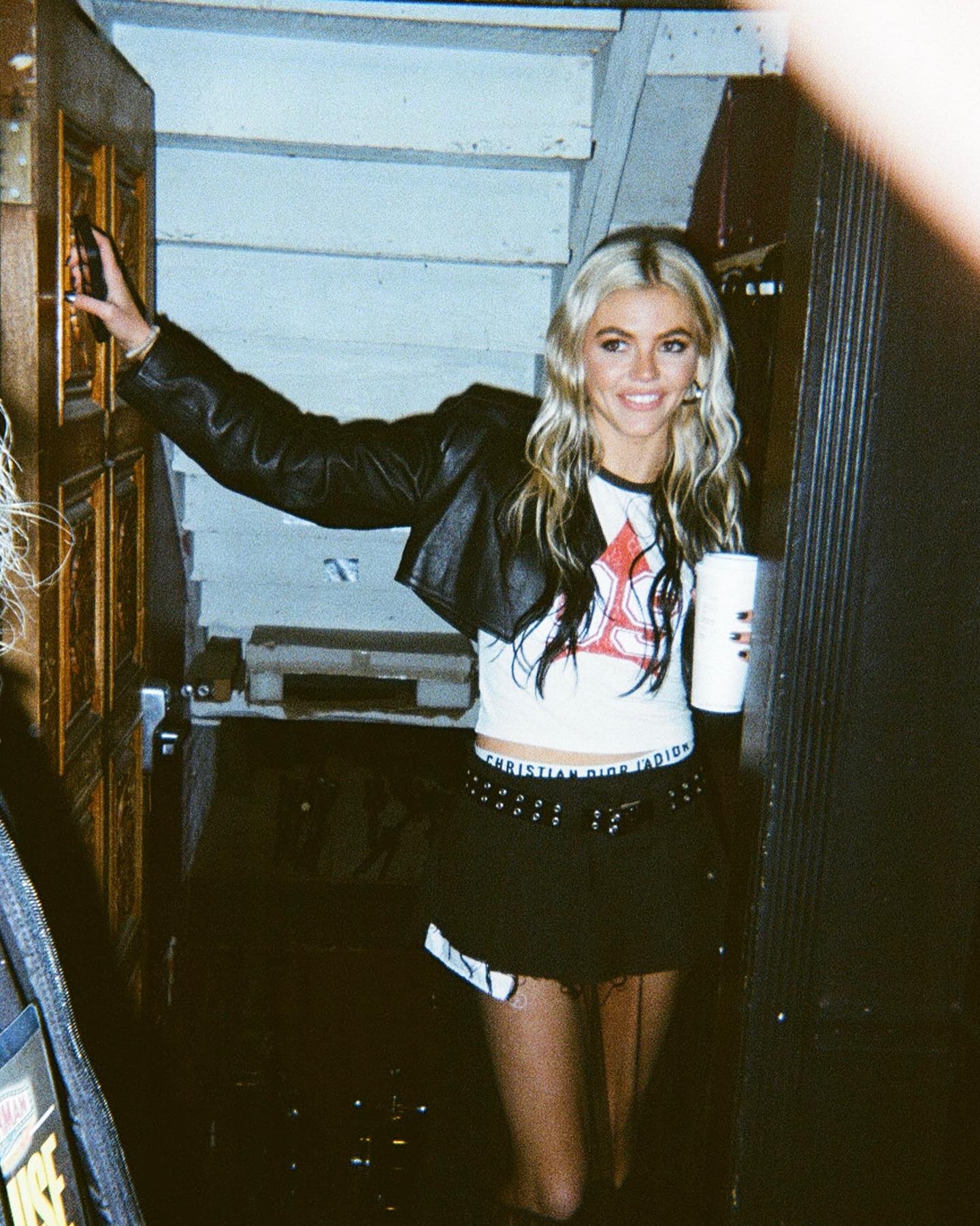 a woman with blond and black hair is smiling while standing in a doorway. she is wearing a black jacket, a white top, and a black mini skirt with a dior logo on the waistband. she is holding a white cup in her right hand and has her left hand on the door frame. the background is a dark interior with stairs visible behind her.