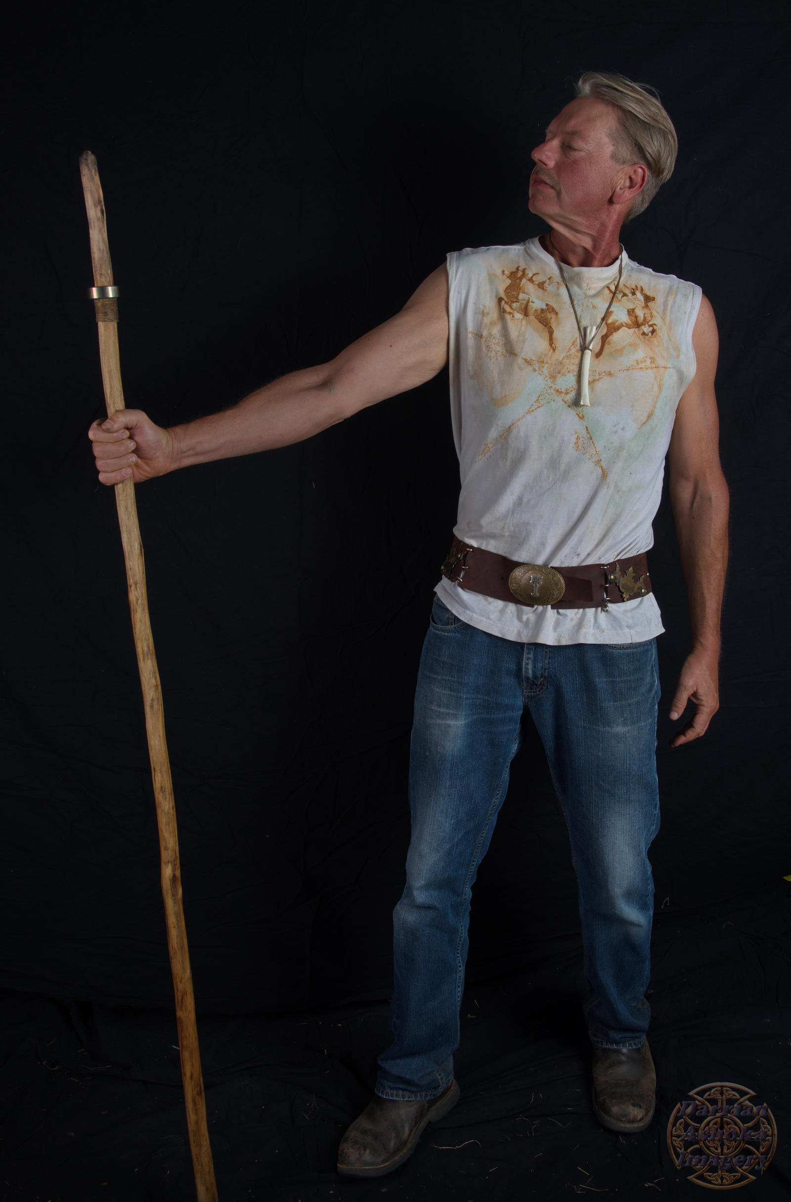 a fullbody studio shot of a man standing against a black background. he is holding a long, rustic wooden staff in his right hand, his arm extended to the side. he is wearing a white sleeveless tshirt with brown stains, blue jeans, and brown leather belt with a large buckle. he also wears a necklace with a bone pendant and brown boots. his expression is serious and he is looking slightly away from the camera.  the overall style is casual, simple, and slightly rustic.