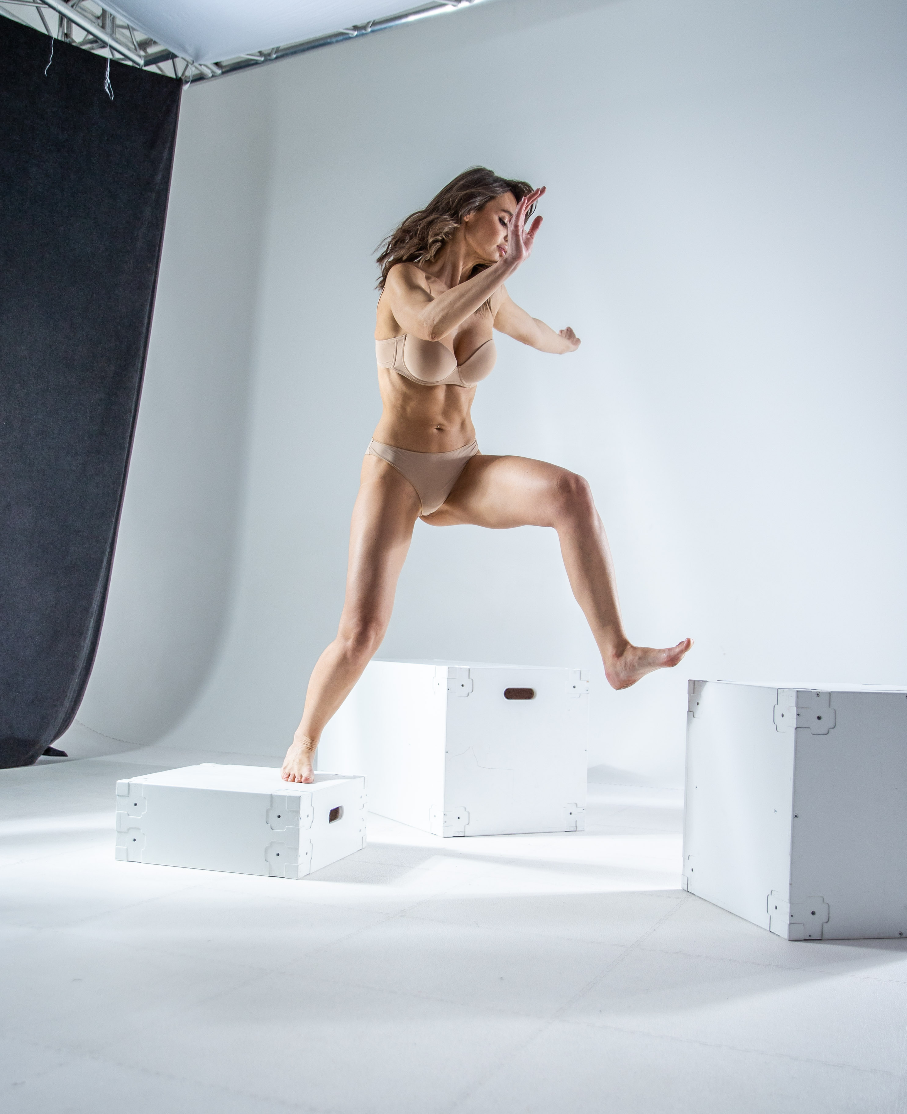 a woman in underwear is jumping from one white box to another in a studio setting. she is barefoot and appears to be in good physical condition. her arms are extended for balance. a black backdrop is visible in the background.