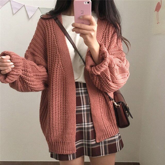 a woman is taking a selfie. she is wearing a pink knit cardigan sweater, a white tshirt, and a plaid skirt. she also has a brown purse slung over her shoulder. the background is a simple white wall with some decorations. the overall style is casual and fashionable.