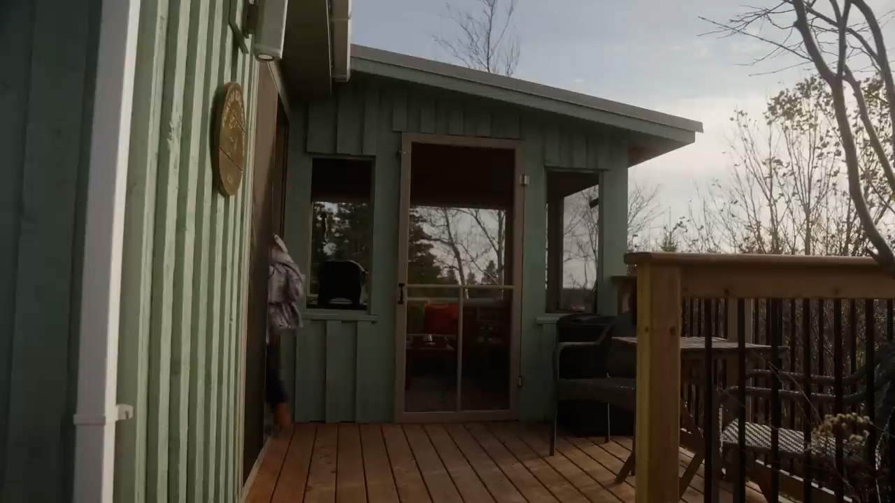 the image shows a person stepping out of a light green wooden cabin onto a wooden deck. the deck is attached to a screenedin porch. the porch has large windows offering a view of the surrounding woods. the trees in the background suggest it might be autumn. the lighting suggests a sunny day with the sun somewhat low in the sky, casting shadows on the deck. the overall feel of the image is peaceful and inviting.