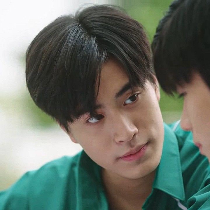 a closeup image featuring two young men, one looking intently at the other with a soft expression on his face. they appear to be wearing matching green uniforms, possibly indicating a school or sports team setting. the background is blurred, emphasizing the connection between the subjects. the lighting is soft and natural.