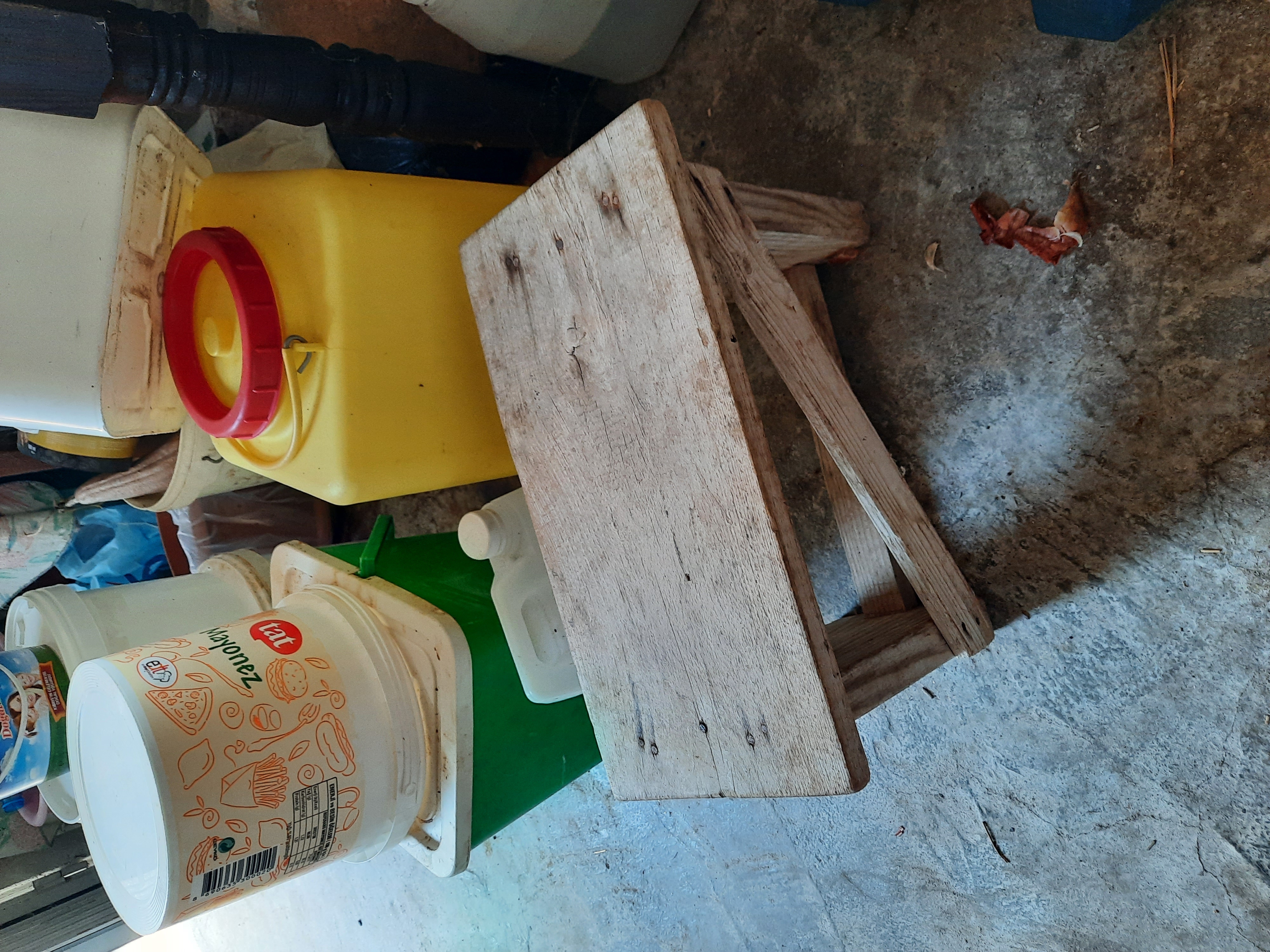 the image shows a cluttered area with a small wooden stool in the foreground. behind the stool are several plastic containers and buckets of various colors and sizes. the floor is made of concrete and appears to be dirty.