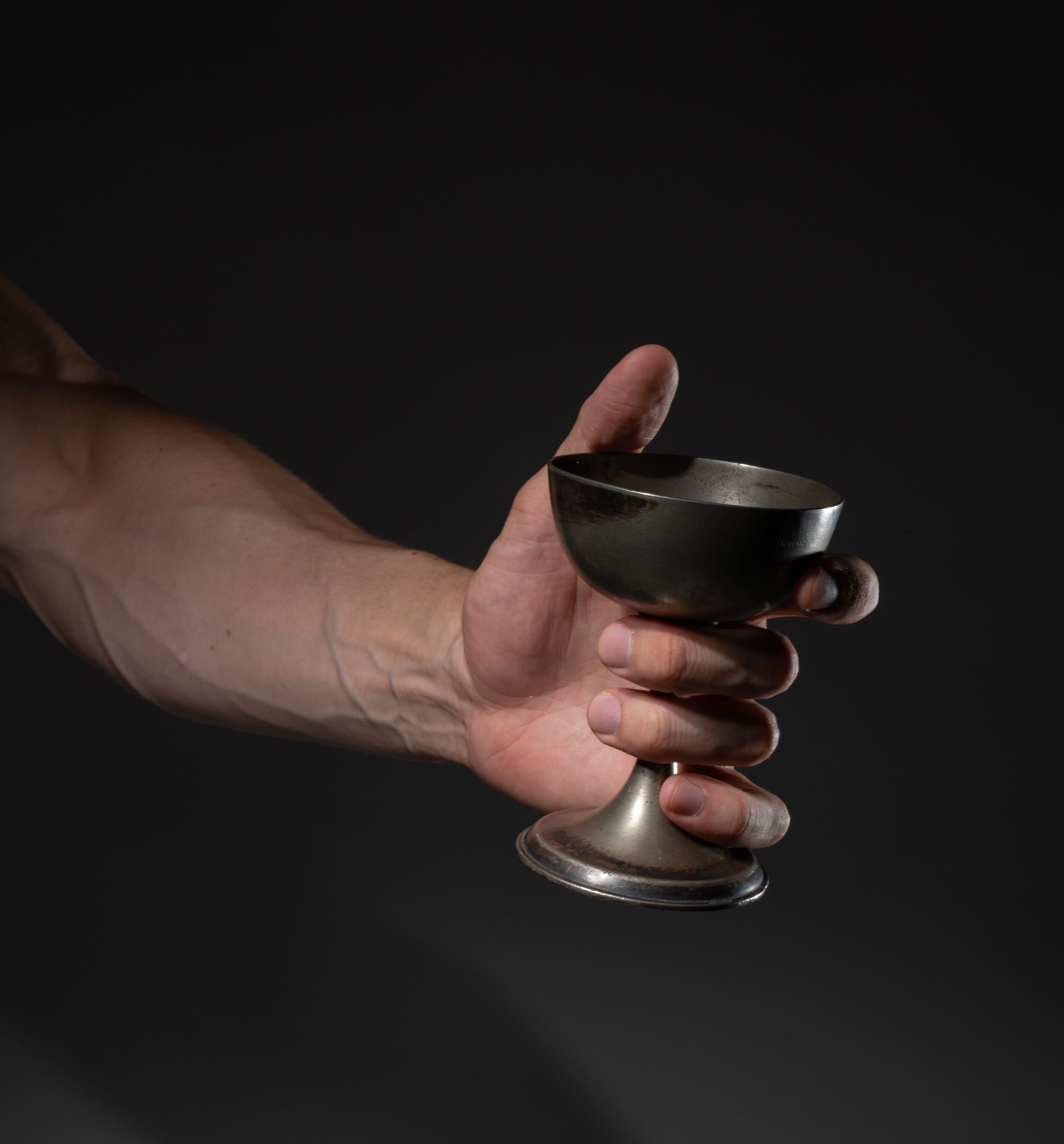 a man's arm extends from the left side of the image, his hand grasping a vintage, slightly tarnished silver cup. the background is a dark gray, and the cup is lit dramatically from the front.
