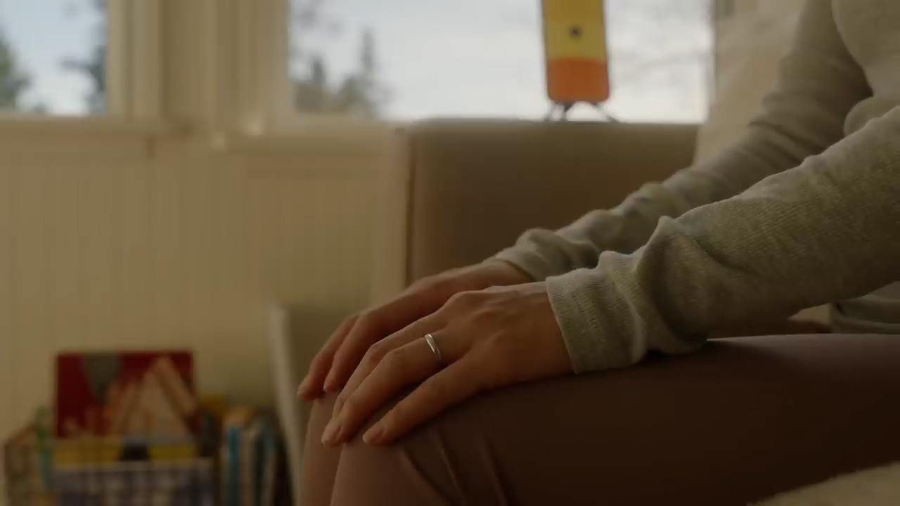 the image shows a closeup of a woman's hands resting on her lap. she is sitting on a couch, and the soft, natural light from a window illuminates the scene. the warm, muted color palette contributes to the overall feeling of peace and quiet.