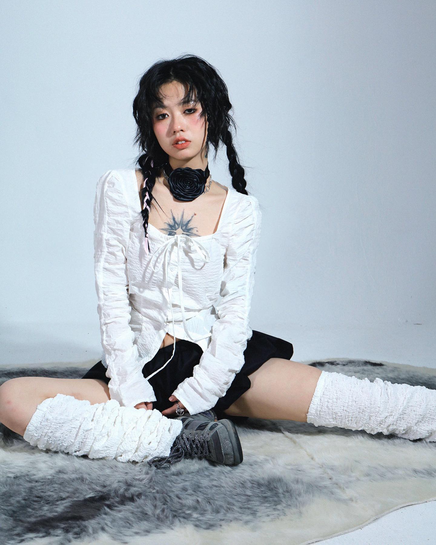 the image shows a young asian woman, possibly a teenager, modeling a stylish outfit. she is sitting on a fluffy white rug against a white background.  she's wearing a white, ruffled, squarenecked blouse with long sleeves, a black mini skirt, and white crocheted leg warmers.  she also has on darkcolored boots. her accessories include a black flower choker and her hair is styled in two braids. she has a small sun tattoo visible on her chest.  the overall style is a mix of alternative, grunge, and possibly dark academia elements. the image is a professional studio shot, likely for a fashion publication or online store.