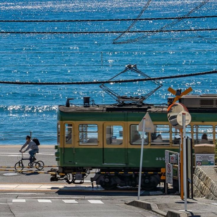 a green and yellow tram is crossing a road near a beach. a person is riding a bicycle in the foreground. the ocean is a bright blue and the sun is shining. the scene is peaceful and picturesque. the tram is number 355.