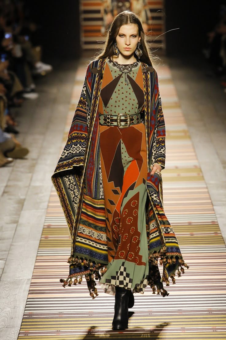 a model is walking on a runway wearing a long, patterned dress, a shawl, a belt, boots, and jewelry. the dress features a variety of geometric patterns and colors, and the shawl has tassels. the model has long hair and is making eye contact with the camera. the runway is decorated with a striped pattern.
