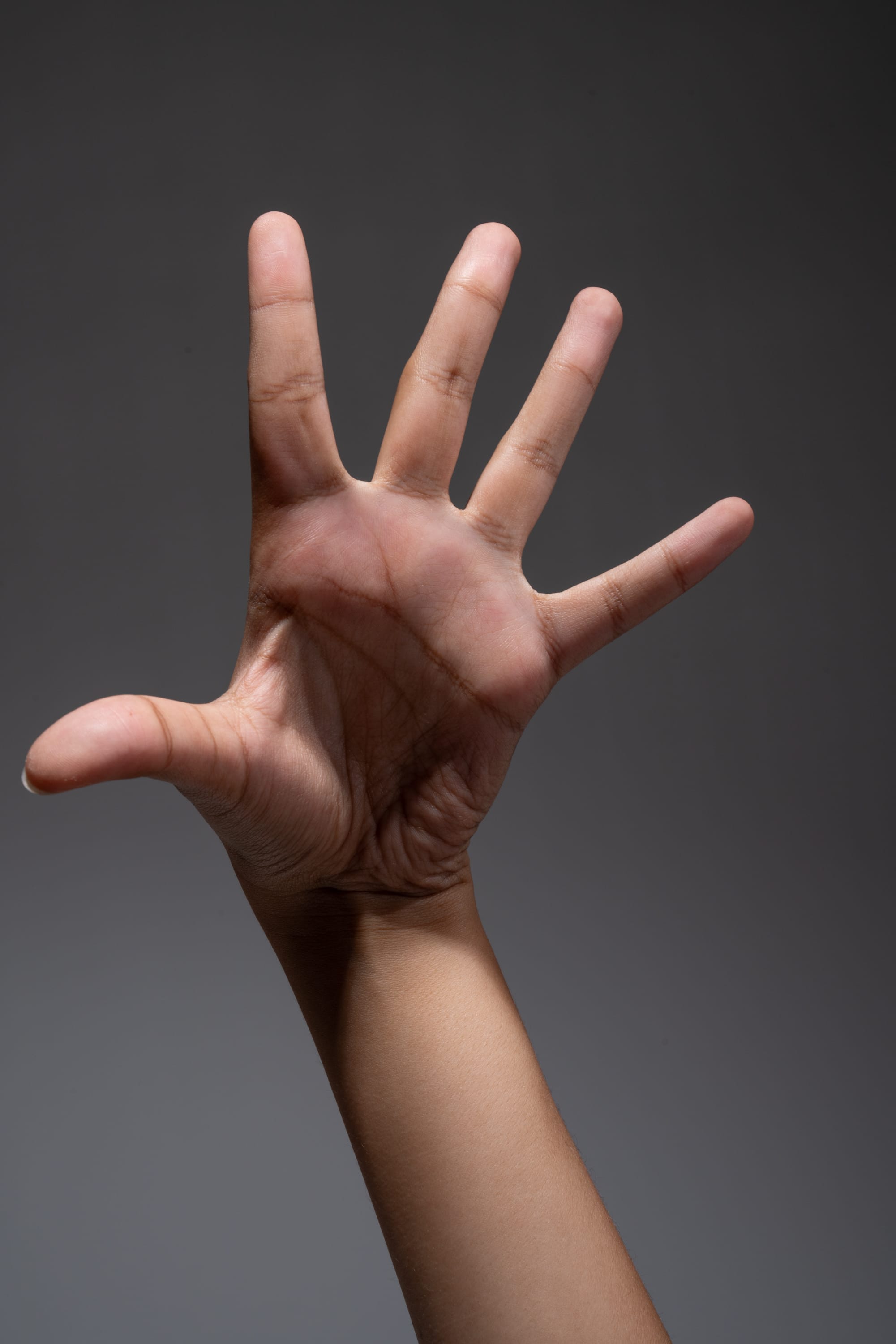 a hand is extended with an open palm, displaying all five fingers, set against a neutral gray background. the lighting suggests a controlled studio environment, highlighting the details of the hand and emphasizing the gesture.