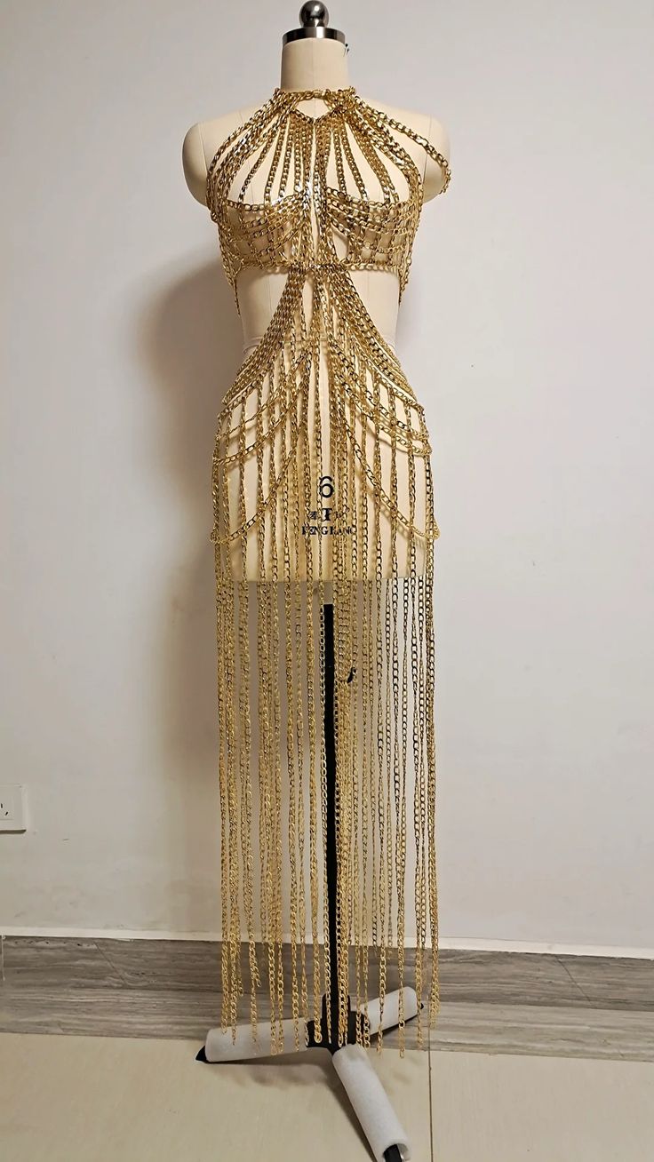 a golden chain dress displayed on a mannequin. the dress is made entirely of golden metal chains, with a halter neckline and a draped skirt. the mannequin is standing in front of a white wall.