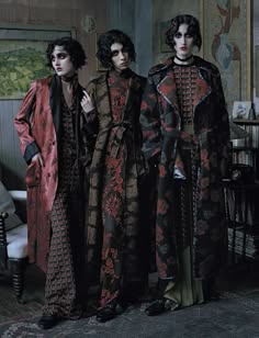 three women dressed in dark, antique, long robes stand in a dimly lit room posing for a portrait. they are looking at the camera, their expressions serious. the women are wearing black and red robes with historical style embellishments and trim. their dark hair is sleek and stylish and their makeup is dramatic and dark. the room is dark and mysterious, and there are antique objects in the background. the overall tone of the image is gothic and vintage, with a sense of mystery and formality.
