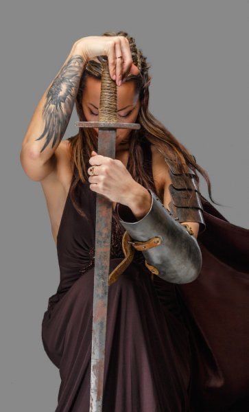 a fierce woman warrior dressed in a maroon dress and armor, grips a medieval sword and stares intently at the camera. the setting is simple and the focus is on the woman's intensity and the danger that she poses.