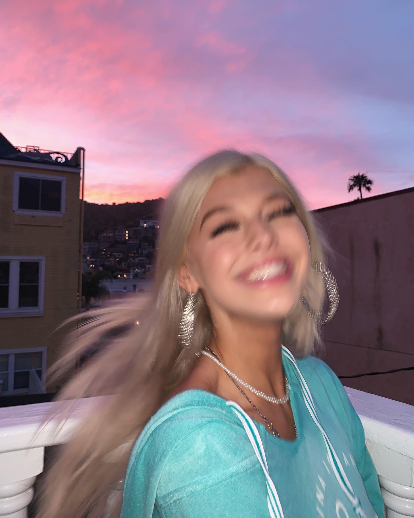 a woman with blonde hair smiles in front of a colorful sunset. she is wearing jewelry, including earrings and a necklace, and a blue sweatshirt. in the background, there are buildings and a palm tree.