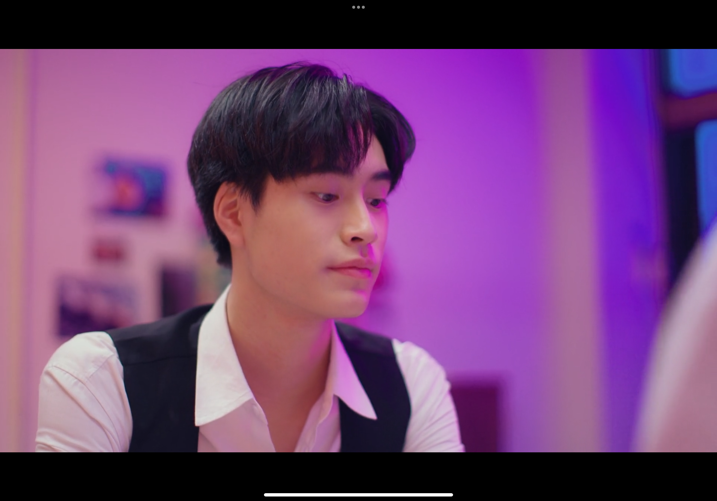 a closeup shot of a young asian man with dark hair. he is wearing a white collared shirt and a black vest. the lighting is predominantly purple and pink neon, creating a moody atmosphere. the man's expression is serious and thoughtful as he looks slightly down and to the right. the background is out of focus, suggesting an indoor setting, possibly a room.