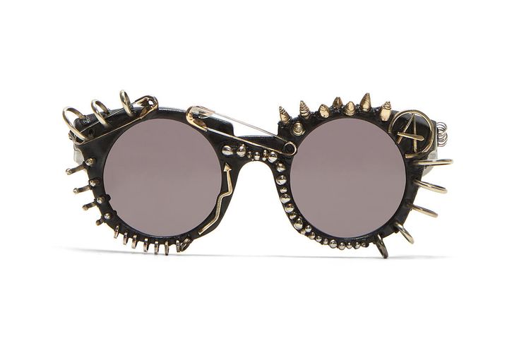 a pair of sunglasses with a punk rock aesthetic. the round lenses are gray and surrounded by a black frame. the frame is heavily embellished with goldcolored studs, spikes, and metal accents. a safety pin is placed across the top of the frames. the right lens has an anarchy symbol on the side.