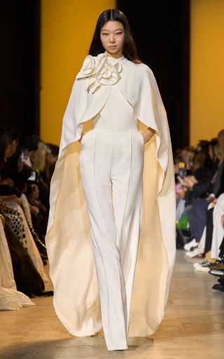 a fashion model walks down a runway, wearing a white pantsuit with a capelike top and a large flower detail. the outfit is very elegant and stylish, and the model looks confident and beautiful. she is wearing a bold makeup look and has her hair pulled back in a sleek style. the image is likely from a high fashion runway show.
