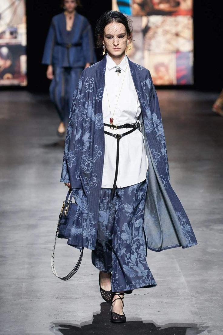 a fashion model walks down the runway wearing a blue denim dress with floral pattern. the dress has a long flowing design and features a white shirt underneath. she is accessorized with a black belt, a small blue bag, and black shoes.  her hair is pulled back and she is wearing a black headpiece and simple jewelry. the runway is a gray concrete surface.