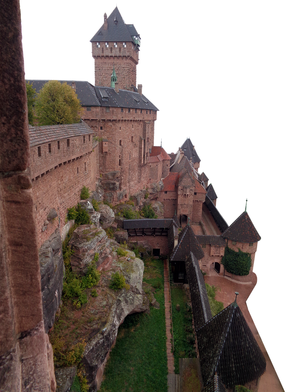 the image shows a view of the château du hautkœnigsbourg castle, a medieval fortress located in alsace, france. the castle's red sandstone walls and towers rise dramatically from a rocky outcrop. the architecture features a mix of romanesque and gothic styles. a tower dominates the view, along with defensive walls, smaller turrets and rooftops. vegetation clings to the rocks below the castle, and grass covers the surrounding area. the sky is blacked out in the background.