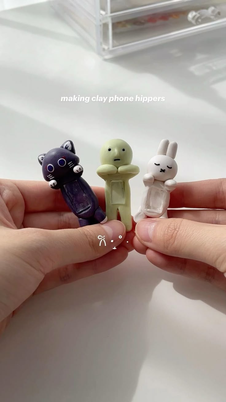 a pair of hands are holding three clay figurines. the figurines are a purple cat, a green human figure, and a white bunny. the text "making clay phone hippers" is visible at the top of the image.