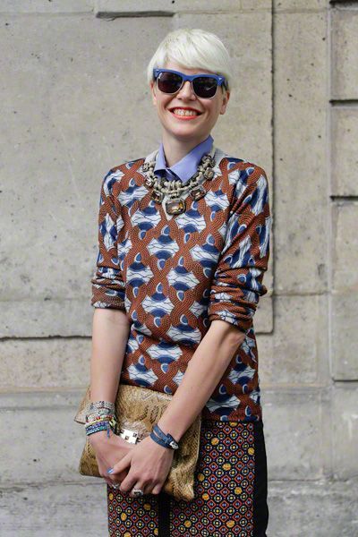 the image showcases a stylish blonde woman with a short pixie haircut, wearing blue sunglasses. she's dressed in a brown and blue geometric patterned sweater paired with a similarly patterned skirt.  she accessorizes with a layered statement necklace, multiple bracelets, and a snakeskin clutch. the backdrop is a stone wall, suggesting an outdoor, street style setting.  her overall look is casual yet chic and fashionable.
