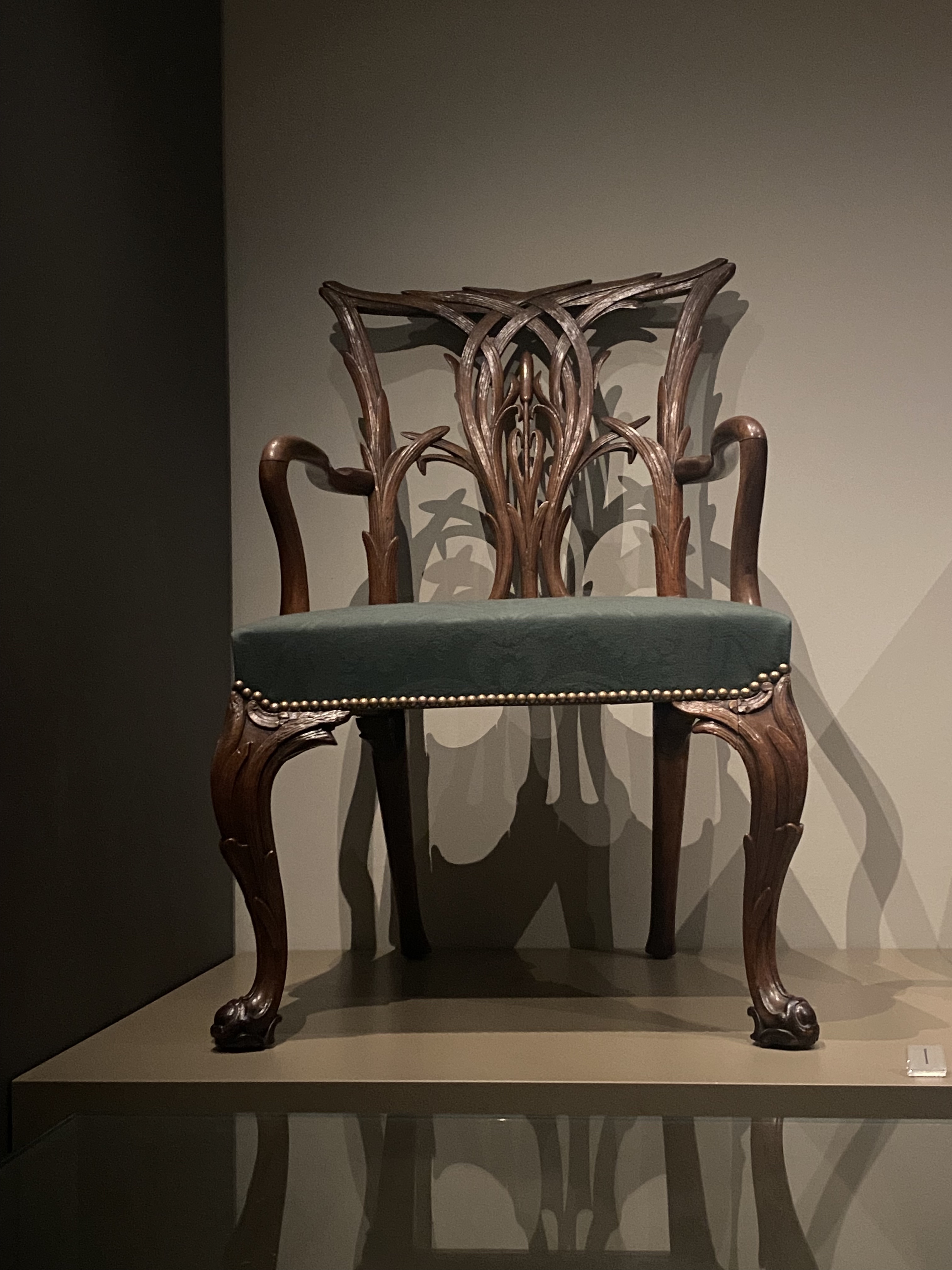 a brown antique chair with an upholstered seat is displayed. the chair is made of wood with ornate, carved details on the back, legs, and armrests. the seat is upholstered in a dark green fabric. the chair is on display in an exhibition setting, standing on a platform against a neutralcolored wall.