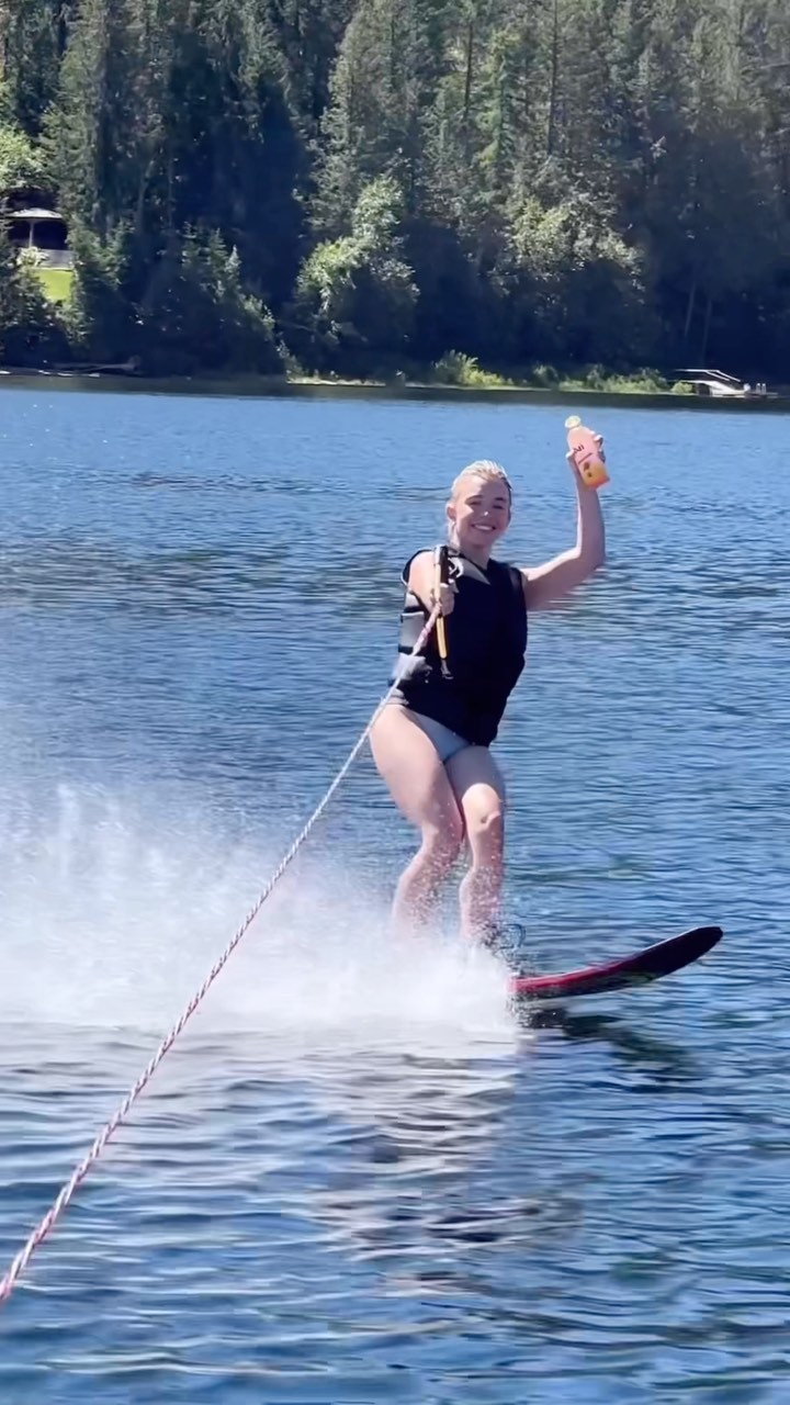 a woman is waterskiing on a calm lake. she is smiling and holding a drink in her hand.  the lake is surrounded by lush green trees. the scene is bright and sunny, portraying a sense of summer fun and outdoor recreation. she appears happy and athletic.