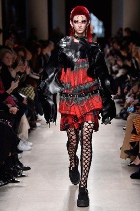a female model walks the runway during a fashion show. she is wearing a red hair, black leather jacket, a plaid skirt, fishnet stockings, boots, and accessories. the model is posing for the cameras and the lighting is bright. the image captures a moment of high fashion glamour.