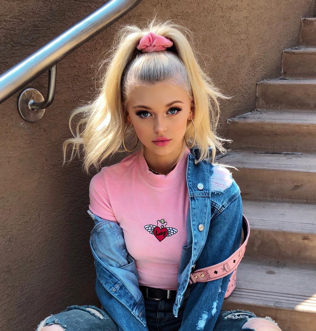 a blonde woman with blue eyes is sitting on stairs, posing for a photo. she is wearing a pink shirt with a heart design, a denim jacket, and hoop earrings. her hair is styled in a high ponytail with a pink hair tie. the image has a selfielike quality, focusing on the woman's face and upper body. she's sitting on a set of stairs, adding depth to the composition. the overall style is fashionable and youthful.