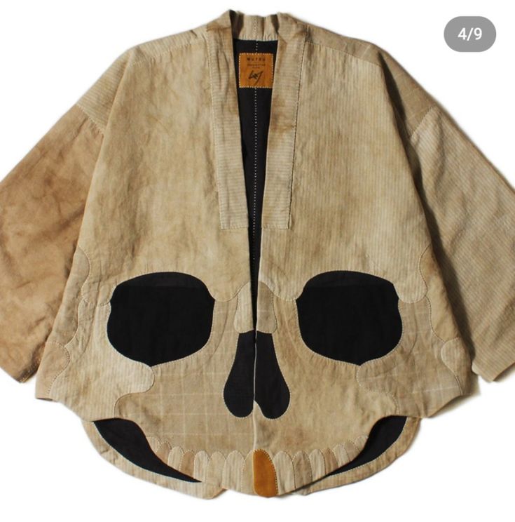 a tan jacket with a large skull design stitched onto the front. it has a wide collar and loose, flowing sleeves. the jacket is made from patchwork material and appears to be vintage or handmade. the skull is made up of black fabric, while the jacket itself is a blend of beige, brown, and tan tones.