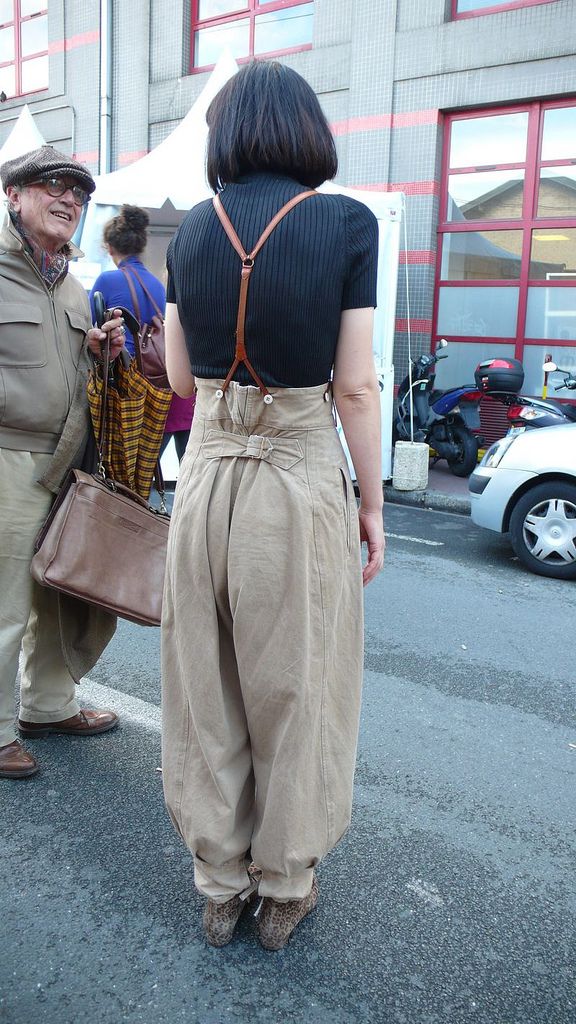 a woman wearing highwaisted pants with suspenders stands on a street, viewed from the back. she has short black hair and is wearing a black top. the pants are beige, and she is also wearing leopard print shoes. a man in a hat and glasses stands beside her. he is wearing a beige jacket and holding a brown leather handbag. there is a vehicle and a building in the background.