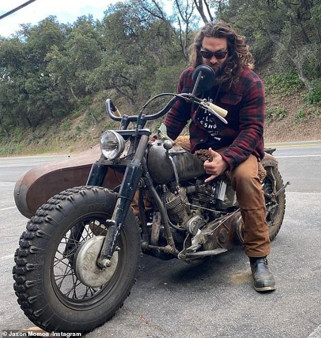 jason momoa sitting on a vintage motorcycle with a sidecar. he is wearing sunglasses, a red and black plaid shirt, brown pants, and boots. he is looking down at the motorcycle. the motorcycle is black and silver and looks old and rusty. the sidecar is brown and rusty. the background is a wooded area and the road is asphalt.