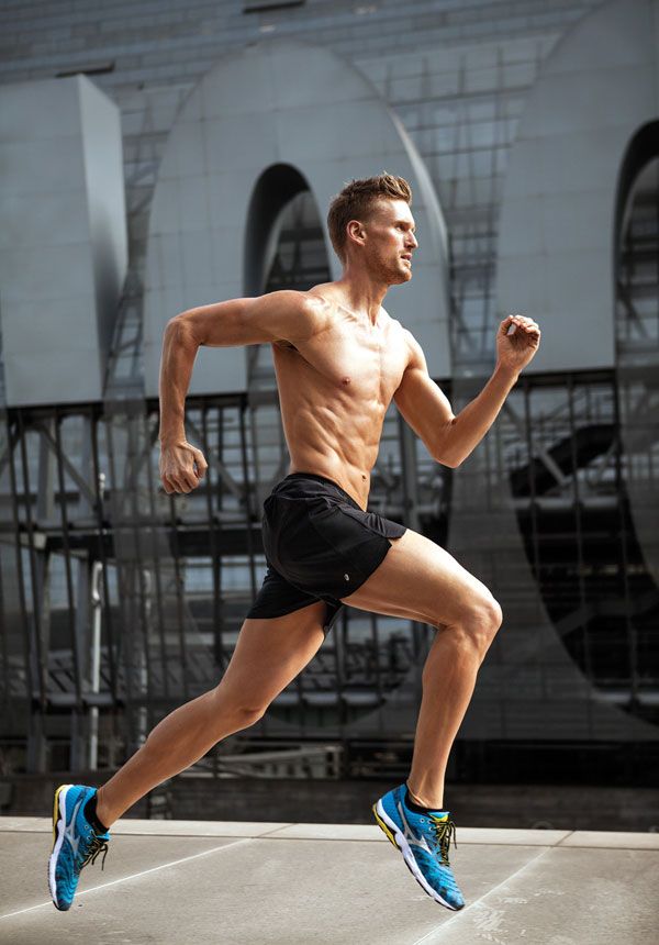 a muscular man wearing black athletic shorts and blue sneakers, running in the city. the man is running toward the camera. the background is a concrete building with a metal structure. the man is looking focused and determined.