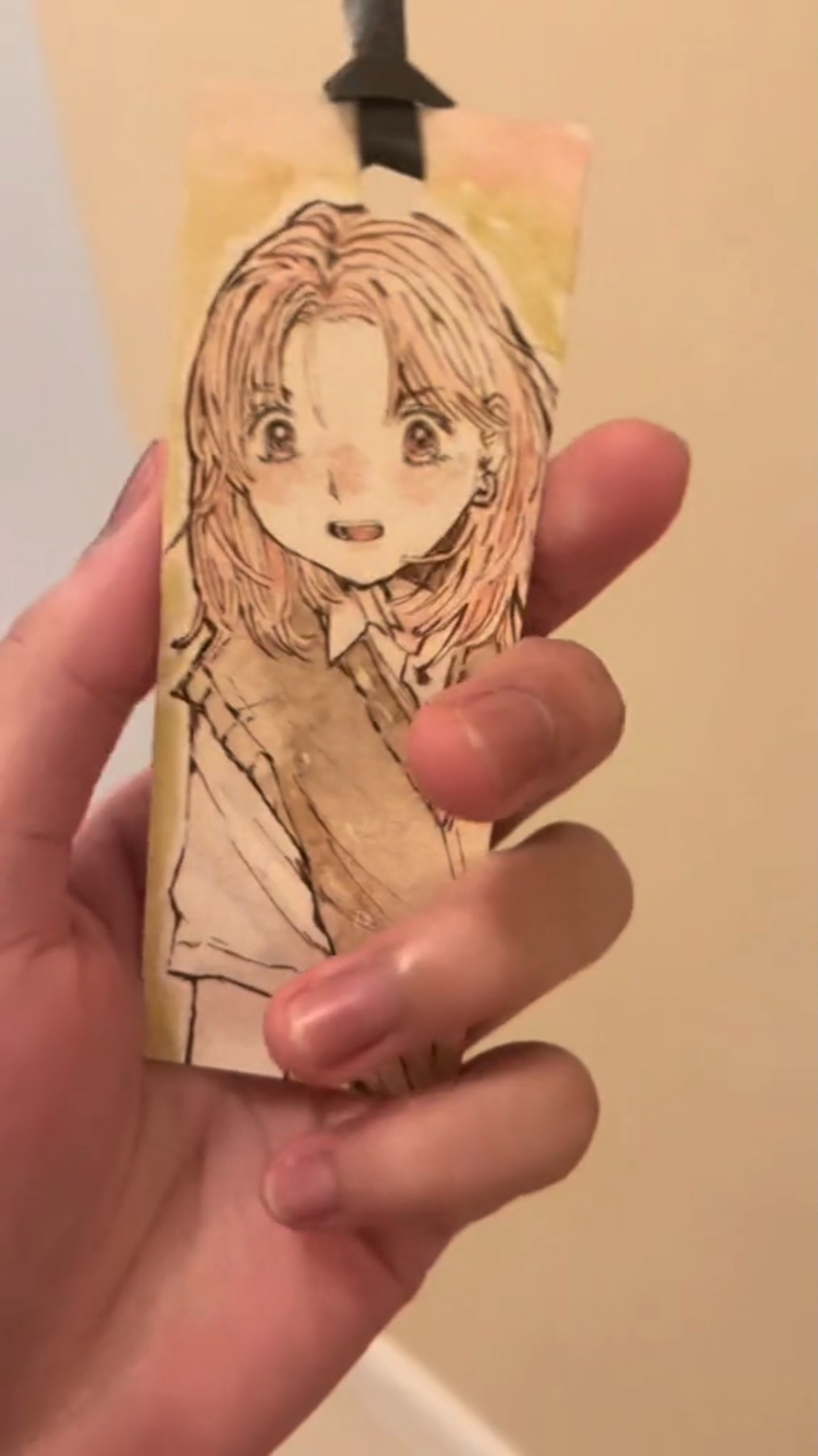 a hand is holding a bookmark with a drawing of an anime girl with long pink hair, drawn in brown and yellow ink on paper. she has a brown jacket and is wearing a white collared shirt. the bookmark is cream colored.