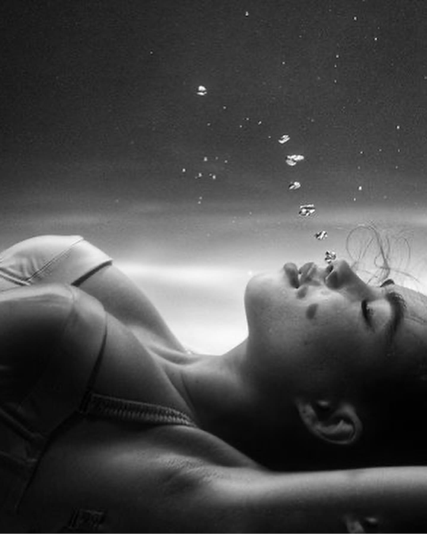 the image is a black and white, closeup underwater portrait of a young woman. she appears serene and relaxed, floating face up in the water.  bubbles rise from below her, creating a soft, ethereal atmosphere. she's wearing a darkcolored bra or top. the overall mood is calm, sensual, and somewhat mysterious. the contrast between the dark water and the woman's light skin enhances the image's dramatic effect.  the focus is on her face and upper body.