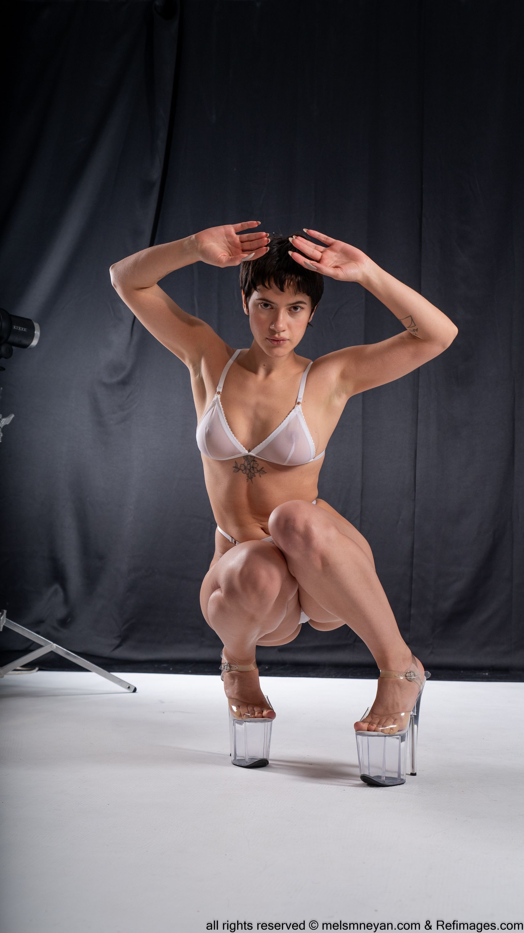 a woman with short hair and tattoos is squatting in a studio wearing a white bra and clear stripper heels. she is posing with her hands up above her head. there is stage lighting equipment to the side of her.