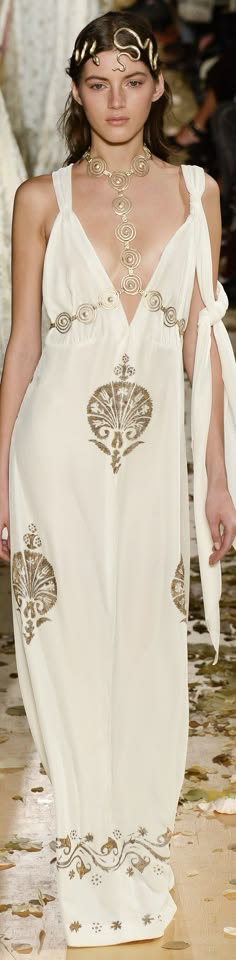 a beautiful woman model is posing on a runway, wearing a white, flowing dress with gold embellishments and a snakelike headpiece. the dress is grecianinspired with a plunging neckline and a flowy skirt. the model's hair is styled in loose waves. the overall look is elegant and beautiful.