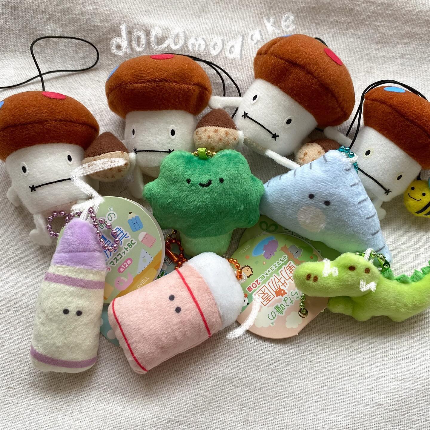 the image shows a collection of plush toys and keychains scattered on a white fabric. there are several mushroom plush toys with brown caps and white stems. among the plush toys, there is a green broccoli plush, a light green crocodile plush, a pink plush, and a blue plush. each plush toy has a keychain attached to it. there are also tags with japanese text on them. the word "docomodake" is embroidered on the top left corner.