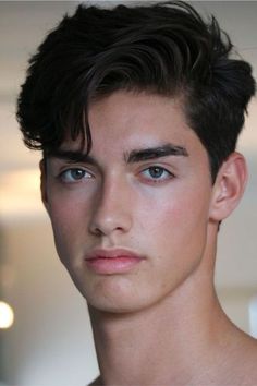 a young man with short, dark brown hair and striking brown eyes is looking directly at the camera. he has fair skin and a neutral expression on his face. the lighting is soft and natural, creating a gentle highlight on his skin. the image is a headshot, cropped at the shoulders. he appears to be indoors.