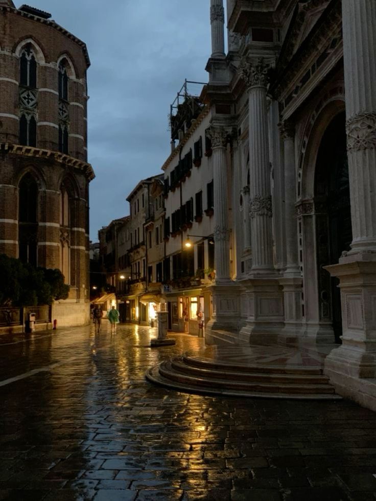 the image captures a captivating night scene in venice, italy.  a rainslicked cobblestone street reflects the soft glow of lamplight, highlighting the intricate details of the historic buildings lining the way.  the architecture is predominantly venetian gothic and renaissance in style.  a grand, columned building dominates the right side of the frame, while on the left, a brick structure, possibly a church or campanile, rises in the background.  the overall atmosphere is moody and atmospheric, with the dampness and low light creating a sense of mystery and quiet beauty.  two figures are faintly visible in the distance, adding to the sense of scale and solitude. the image's composition effectively uses leading lines and reflections to draw the viewer's eye through the scene.