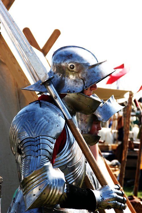 the image showcases a knight clad in shining armor, standing in profile against a blurred background of a medieval reenactment event. the sunlight glints off the polished metal of his helmet, breastplate, and gauntlets. he holds a long spear, its point angled towards the ground. the knight appears to be in his element, embodying the spirit of a bygone era of chivalry and combat.