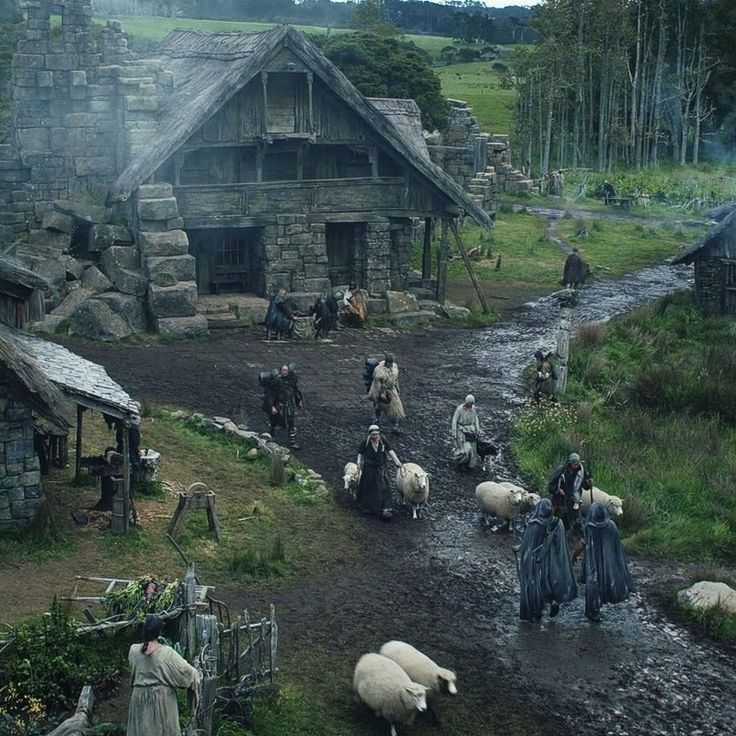 a rustic village scene featuring stone houses with thatched roofs, people in cloaks, and sheep moving along a muddy path. in the background are trees, a forest, and rolling hills. the scene has a medieval or rural aesthetic.
