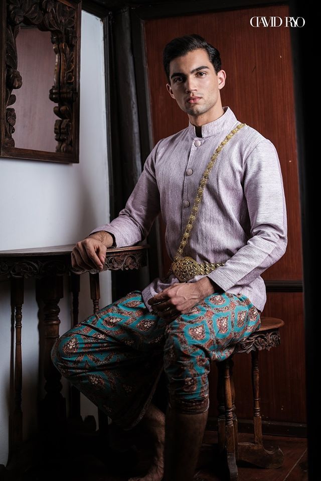 a young man in a traditional thai outfit sits on a wooden chair in an ornate, antique interior. he is wearing a longsleeved, pale pink shirt, teal and gold patterned pants, and a gold belt with a decorative clasp. he has a serious expression on his face, and he is looking directly at the camera. the background of the image is a woodpaneled wall, and there is a mirror on the wall to the left. the man's posture is relaxed, and the image is well lit.