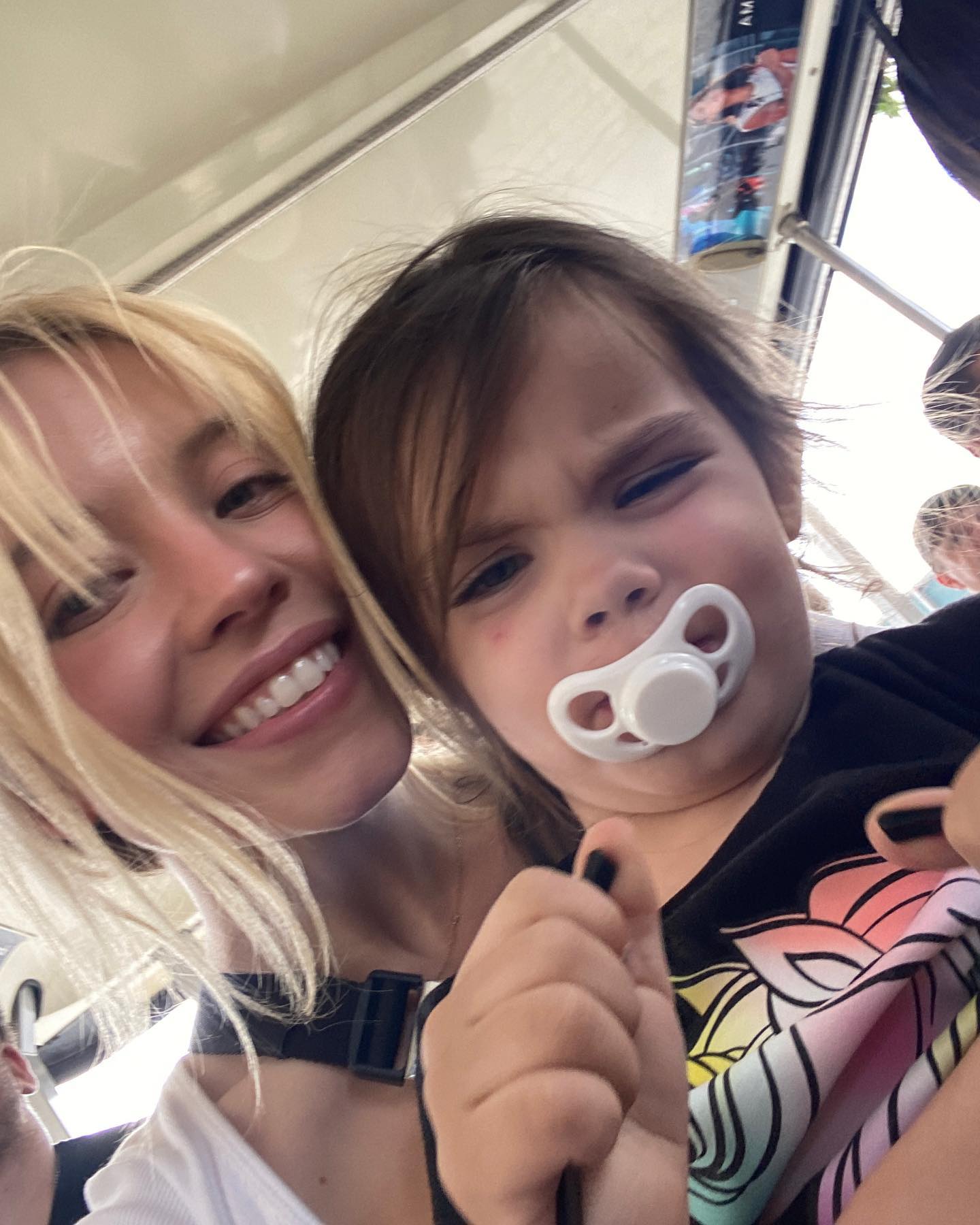 a closeup selfie shows a blonde woman smiling broadly and holding a young girl with brown hair.  the girl is wearing a black shirt with a rainbow design and has a pacifier in her mouth.  they appear to be on a bus or some form of public transport, as parts of the vehicle's interior are visible in the background. the overall mood is warm and affectionate.