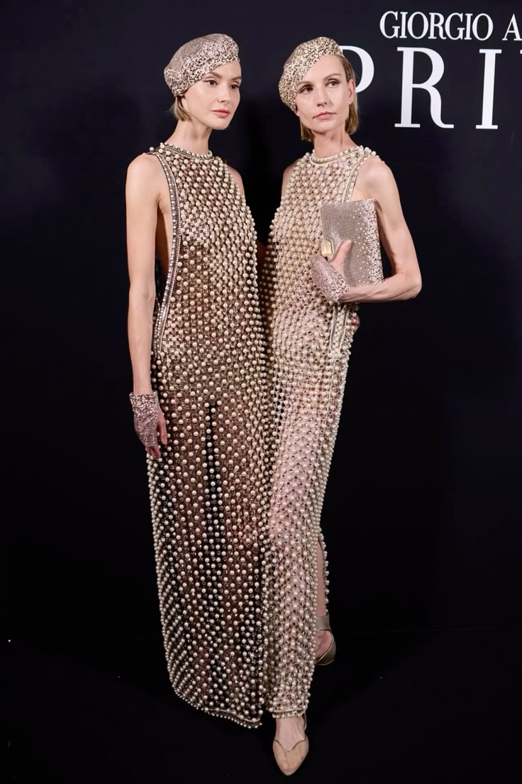 two women are posing on a black background. they are both wearing pearlembellished sheer dresses and matching berets. the woman on the left has a lace wrist cuff and the woman on the right is holding a matching pearl clutch. a "giorgio armani" logo is visible in the background.
