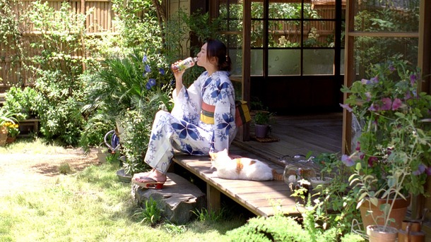 a woman wearing a light blue and white yukata sits on the wooden steps of a traditional japanese house. she is drinking from a bottle. a white and orange cat is lying on the steps next to her. the setting is a beautiful and serene japanese garden with lush green plants, bamboo fencing and flowering plants.  the scene is peaceful and evokes a sense of traditional japanese culture and lifestyle.